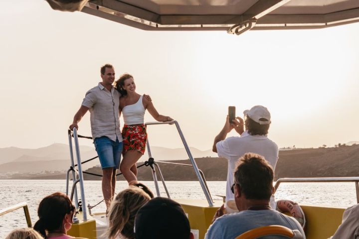 Couple posing on boat at sunset while man takes photo with smartphone.