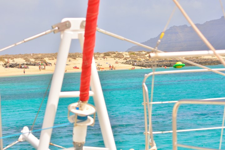 View of sandy beach with people, seen from a sailboat over turquoise water.