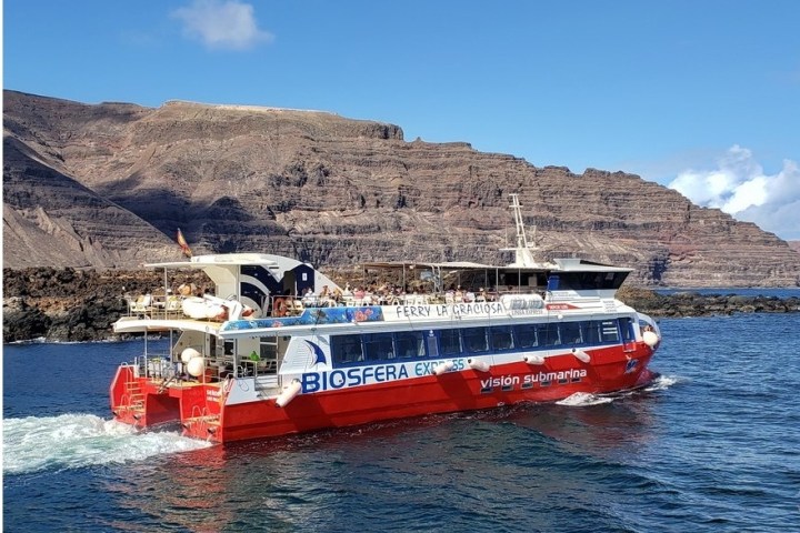 Red and white ferry named 'Biosfera Express' on blue water with rocky cliffs in the background.