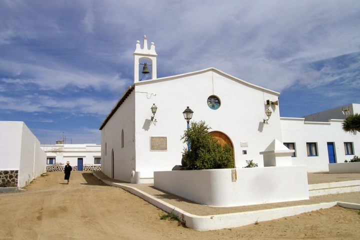 White church with bell tower and arch door, surrounded by sandy path under blue sky.