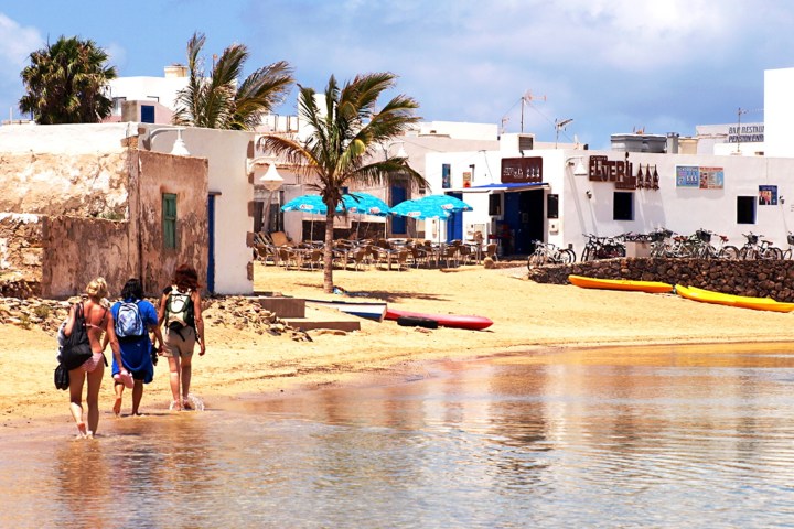 Three people walk along a beach near rustic buildings and palm trees under a blue sky.