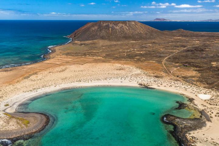 Aerial view of a turquoise bay, sandy beach, and volcanic landscape under blue skies.
