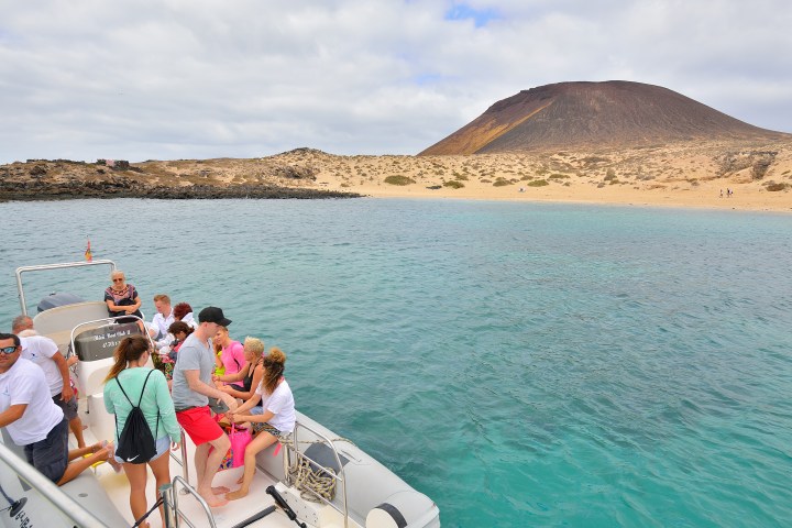 People on a boat near turquoise water and a volcanic island with a sandy coastline.
