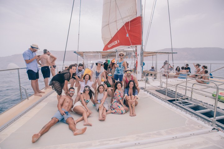 Group of people enjoying a sunny day on a sailboat deck with calm sea and distant mountains.