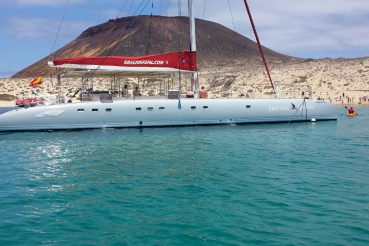 A catamaran anchored near a sandy beach with a volcanic hill in the background.