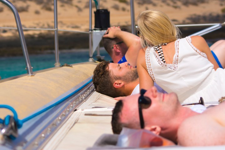People relaxing on a boat deck under the sun near a sandy shoreline.