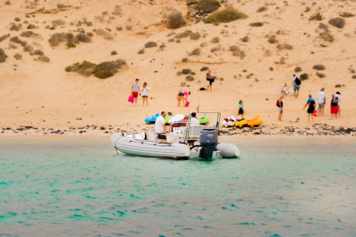 People on a sandy beach with a small boat on turquoise water in the foreground.