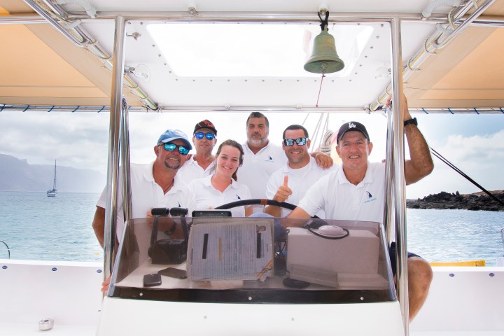 Six people on a boat smiling at the camera with ocean and sailboat in the background.