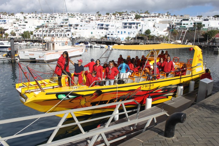 Group in red jackets on a yellow boat docked at a marina with white buildings in the background.