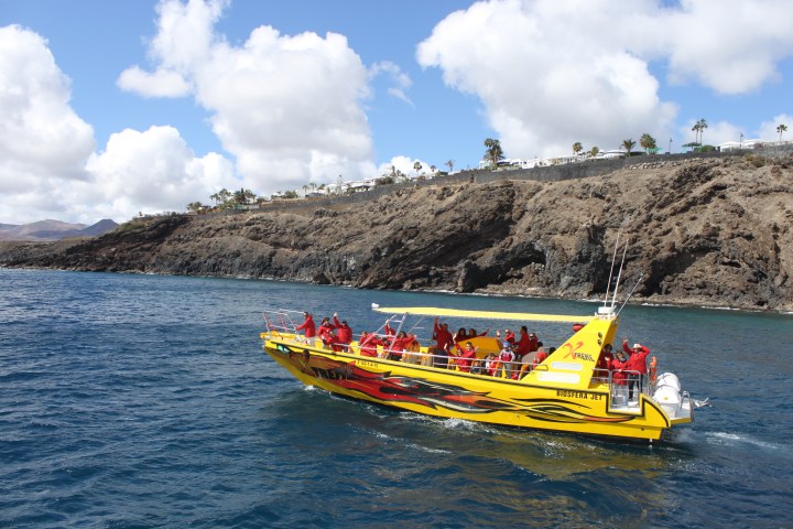 Yellow tour boat with passengers in red jackets near rocky coastline and blue sea.