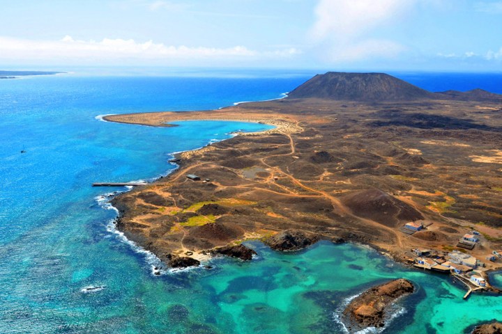 Aerial view of a volcanic island with turquoise waters and rugged terrain under a clear blue sky.