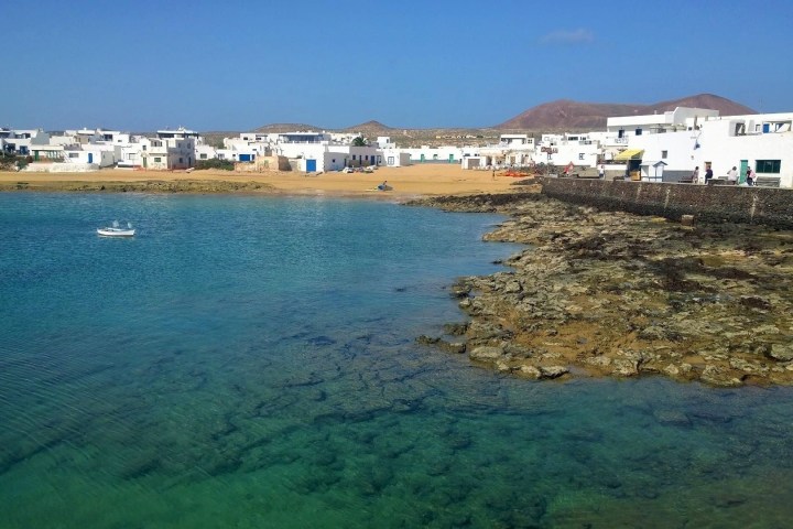 Coastal village with white buildings, sandy beach, and clear turquoise water under a blue sky.