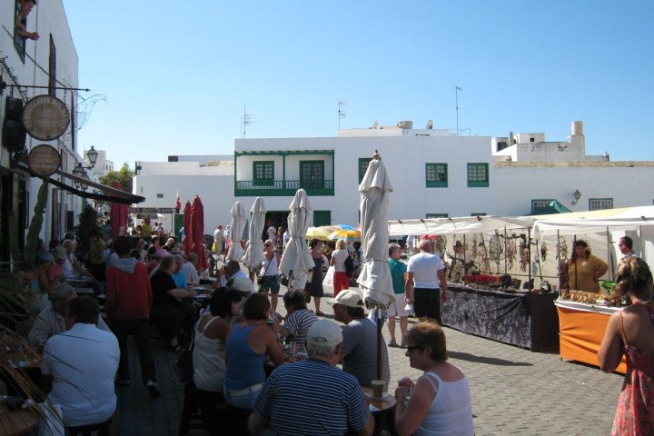 People sitting and shopping at an outdoor market with white buildings and stalls under a clear blue sky.