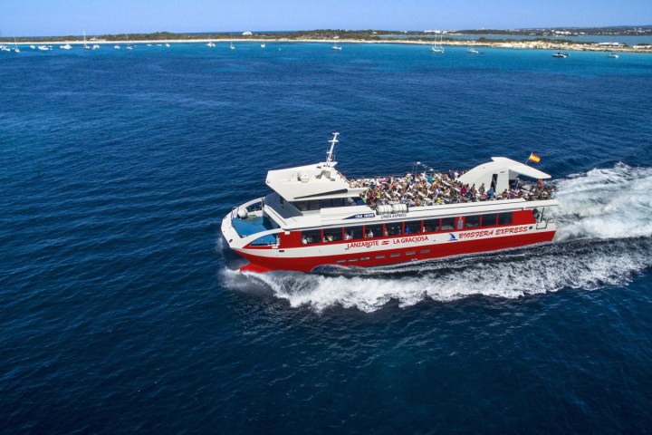 Red and white ferry with passengers sailing on blue water near a distant shoreline.