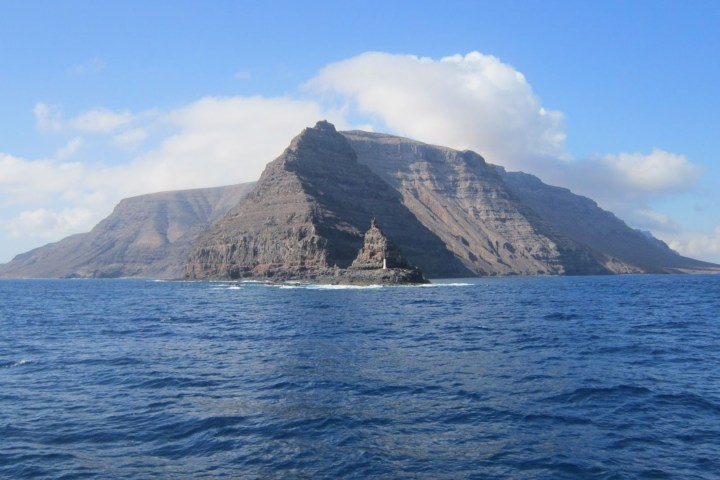 Rocky mountain island with lighthouse surrounded by ocean under a blue sky with clouds.