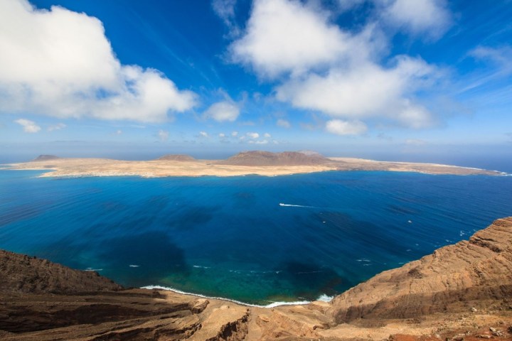 Coastal landscape with a vast blue ocean bay and rocky cliffs under a clear sky with scattered clouds.