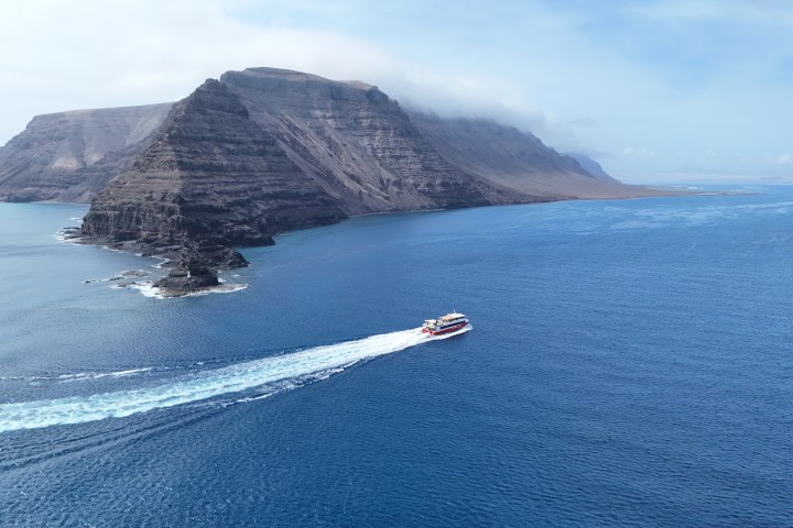 A ferry sails near a rugged coastal cliff under a partly cloudy sky.