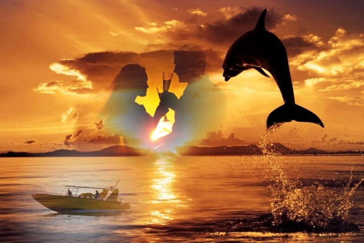Sunset over ocean with couple toasting, dolphin jumping, and small boat on water.