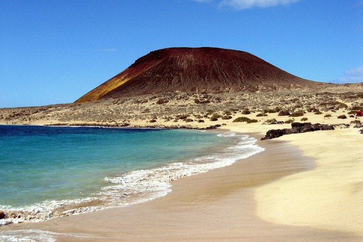 Sandy beach with blue water and a large volcanic hill under a clear blue sky.